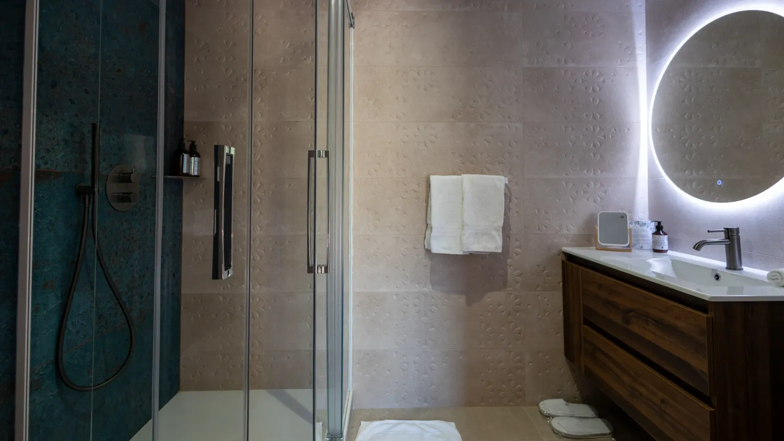 Salle de bain moderne avec douche à paroi en verre, lavabo en bois, miroir rond éclairé et serviettes suspendues