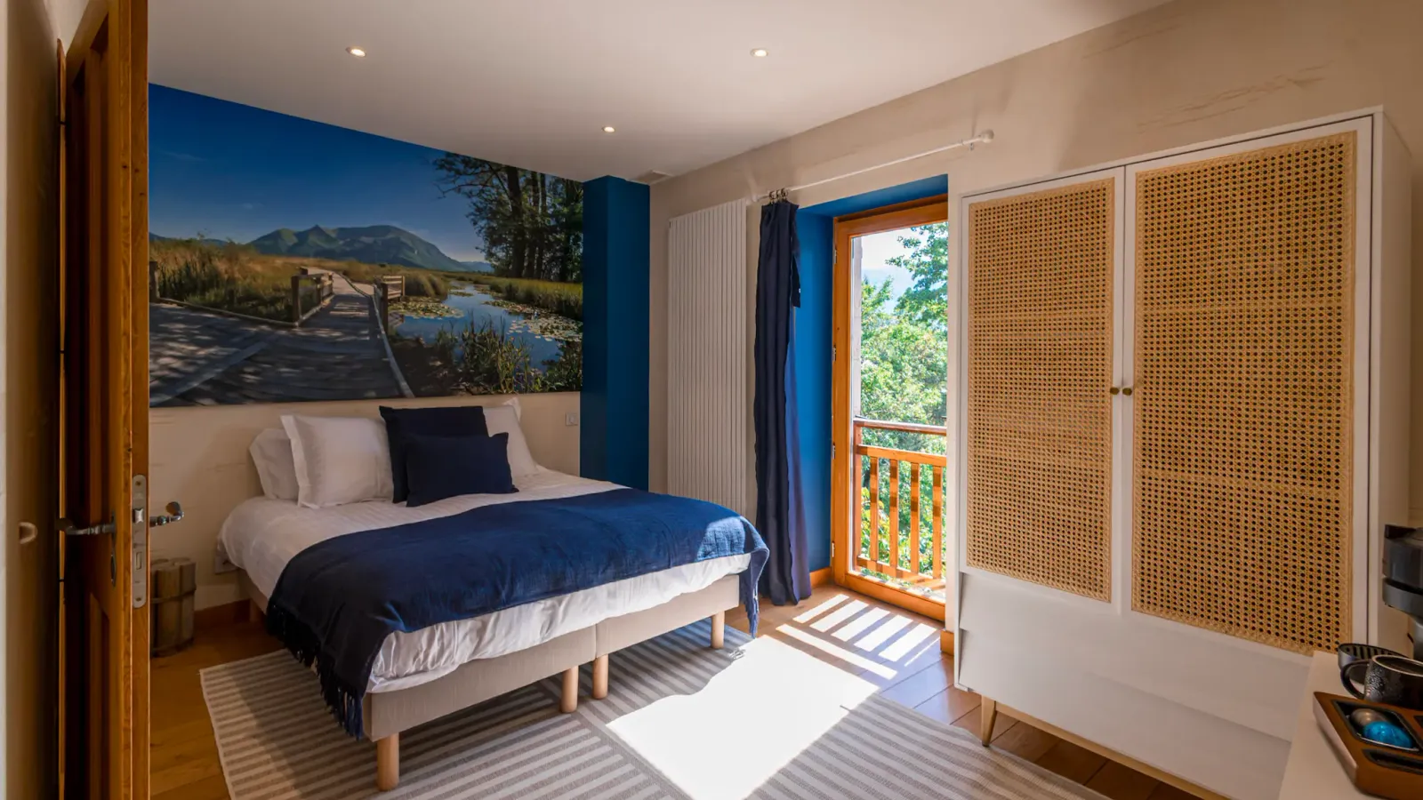 Chambre avec un lit recouvert d'une couverture bleue et blanche, un mur décoré d'une image de paysage naturel, une porte-fenêtre ouverte sur un balcon et un paravent en osier