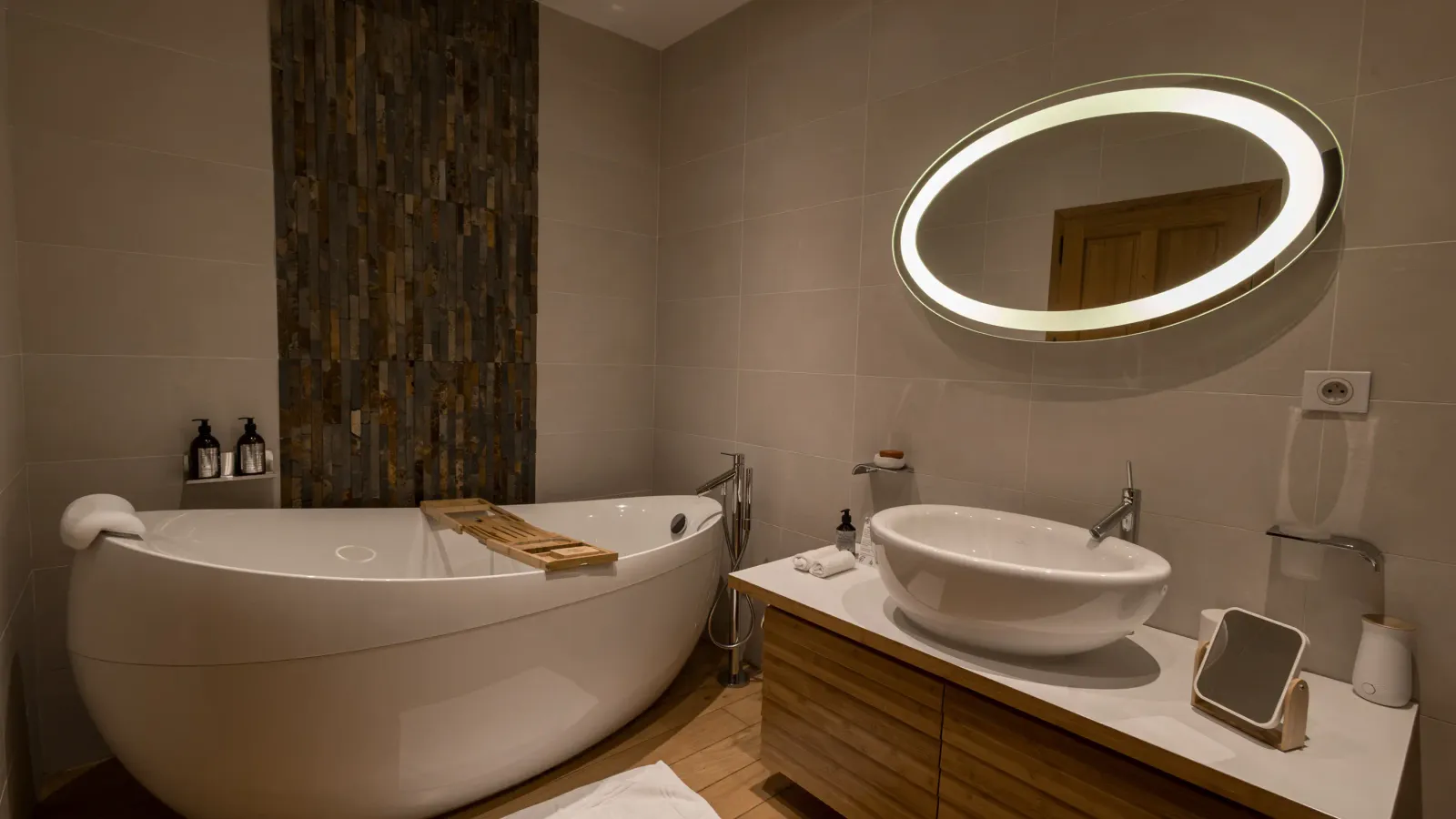 Salle de bain avec baignoire blanche, lavabo et miroir rond éclairé, murs carrelés et sol en bois