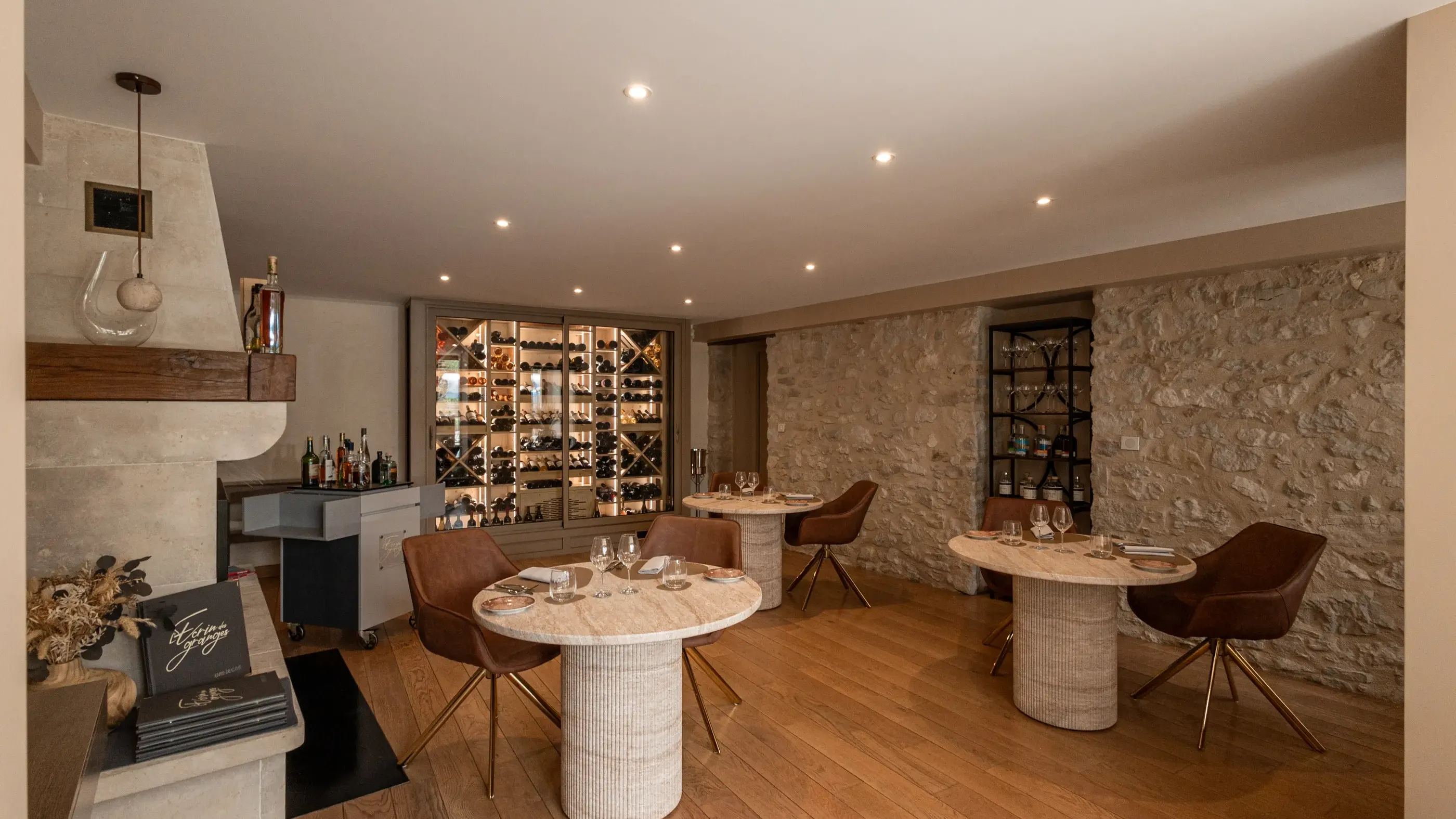 Salle avec tables rondes en marbre, chaises en cuir, mur en pierre, bar avec bouteilles et étagères à vin éclairées